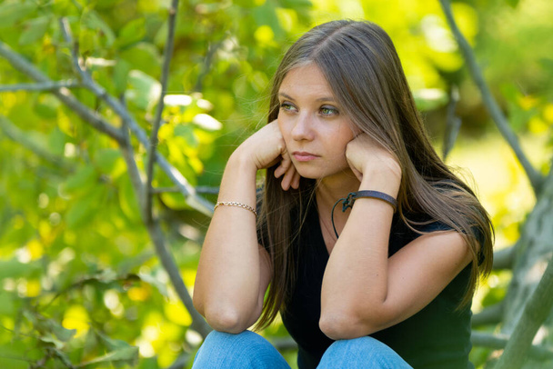 Грустная девушка сидит на фоне листвы и печально смотрит вдаль - Фото, изображение