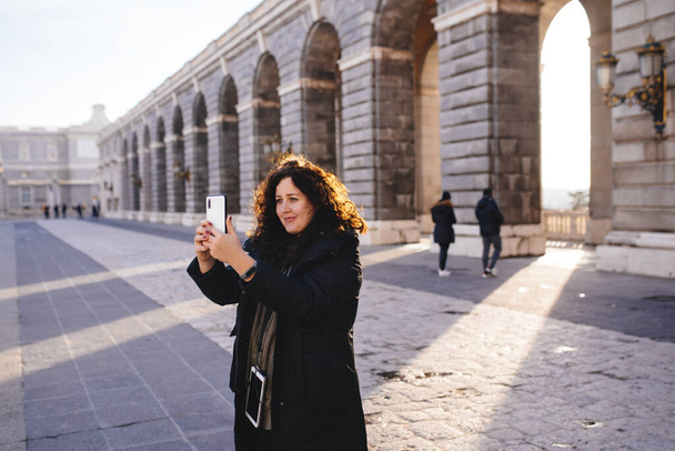 mosolygós lány csinál egy szelfit telefonon egy európai városban, télikabátban, Royal Palace of Madrid - Fotó, kép