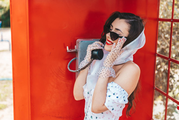 όμορφη νεαρή γυναίκα μιλάει ευτυχισμένα στο τηλέφωνο σε ένα αγγλικό στυλ κόκκινο τηλεφωνικό θάλαμο. Ένα κορίτσι ντυμένο με λευκό φόρεμα και γυαλιά. pinup. - Φωτογραφία, εικόνα