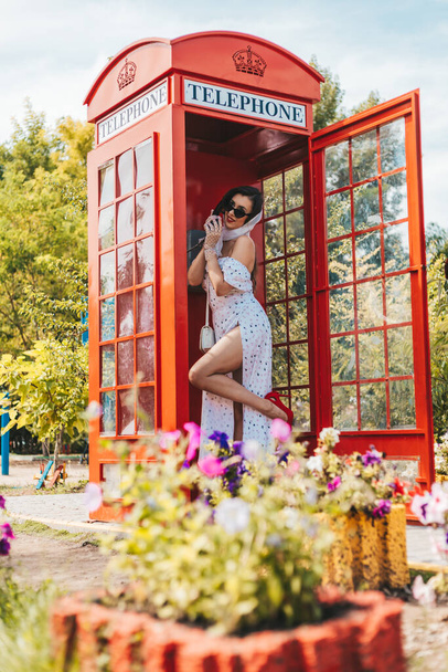 mooie jonge vrouw spreekt graag aan de telefoon in een Engels stijl rode telefooncel. meisje gekleed in een witte jurk en bril. pinup. - Foto, afbeelding