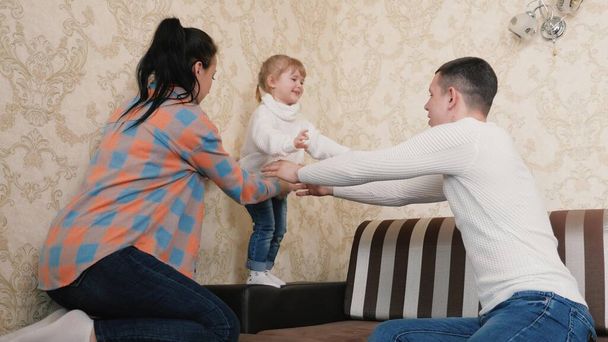 Happy daughter jumps from the couch to dad and hugs him. The childs mother and dad are playing together, having fun at home. Active family vacation at home. Happy childhood, family play at home - 写真・画像