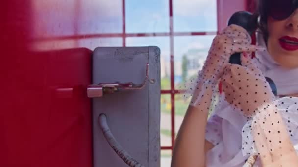 όμορφη νεαρή γυναίκα μιλάει ευτυχισμένα στο τηλέφωνο σε ένα αγγλικό στυλ κόκκινο τηλεφωνικό θάλαμο. Ένα κορίτσι ντυμένο με λευκό φόρεμα και γυαλιά. pinup. - Πλάνα, βίντεο