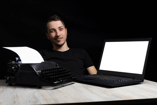Mladý pohledný muž uprostřed mezi psacím strojem a laptopem na černém pozadí v tichosti. Symbol tvořivosti. Koncept minulosti a budoucnosti. Čas a změna - Fotografie, Obrázek