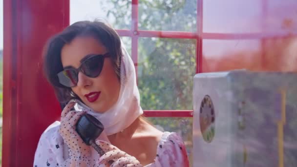Die schöne junge Frau telefoniert fröhlich in einer roten Telefonzelle im englischen Stil. Mädchen in weißem Kleid und Brille. pinup. - Filmmaterial, Video