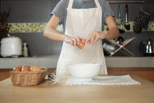 Una cuoca in grembiule bianco sta incrinando un uovo in una tazza per preparare un pasto su un tavolo di legno nella cucina di casa. Mangiare tuorli d'uovo è una colazione sana. - Foto, immagini