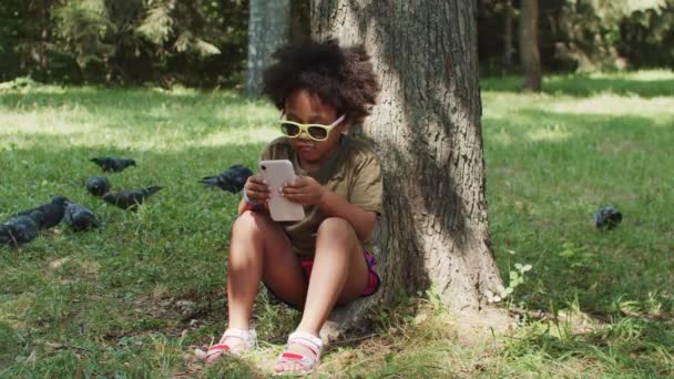 Ένα μικρό μαύρο κορίτσι με γυαλιά ηλίου κάθεται κάτω από το δέντρο και παίζει ένα παιχνίδι στο τηλέφωνο - Πλάνα, βίντεο