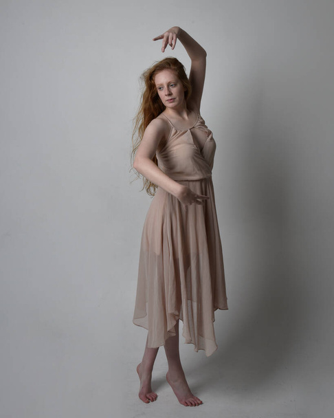Full length portrait of pretty red haired woman dancer,  wearing skin toned flowing fairy dress. Standing gestural poses  isolated on studio background. - Photo, Image