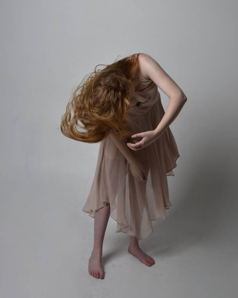 Portrait complet de jolie danseuse aux cheveux roux, vêtue d'une robe de fée fluide et tonique. poses gestuelles debout isolé sur fond de studio. - Photo, image