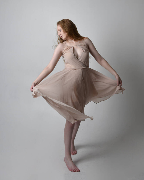 Full length portrait of pretty red haired woman dancer,  wearing skin toned flowing fairy dress. Standing gestural poses  isolated on studio background. - Φωτογραφία, εικόνα