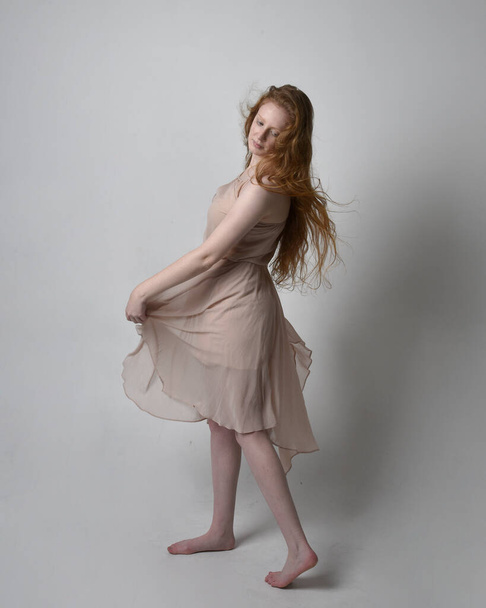 Full length portrait of pretty red haired woman dancer,  wearing skin toned flowing fairy dress. Standing gestural poses  isolated on studio background. - Foto, afbeelding