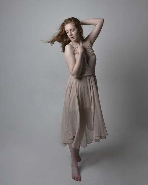 Full length portrait of pretty red haired woman dancer,  wearing skin toned flowing fairy dress. Standing gestural poses  isolated on studio background. - Foto, imagen