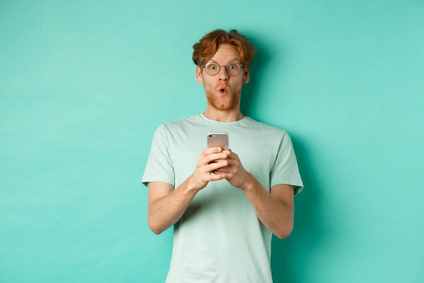 Вражений молодий чоловік з рудим волоссям і окулярами, тримає мобільний телефон, кажучи, що вау і погляд здивовані на камеру, стоїть над м'ятним тлом
 - Фото, зображення