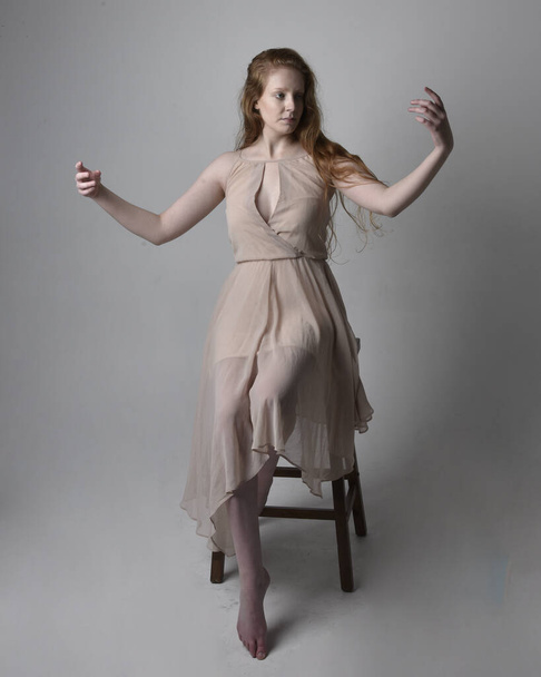 Full length portrait of pretty red haired woman dancer,  wearing skin toned flowing fairy dress. Sitting gestural poses  isolated on studio background. - Фото, зображення