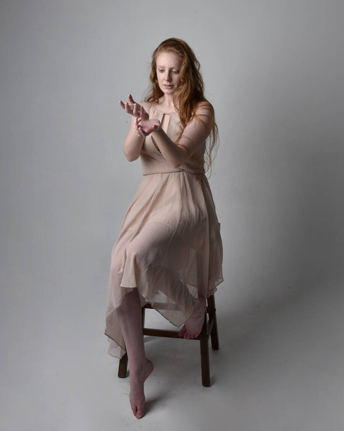 Full length portrait of pretty red haired woman dancer,  wearing skin toned flowing fairy dress. Sitting gestural poses  isolated on studio background. - Φωτογραφία, εικόνα