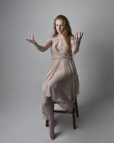 Full length portrait of pretty red haired woman dancer,  wearing skin toned flowing fairy dress. Sitting gestural poses  isolated on studio background. - Fotó, kép