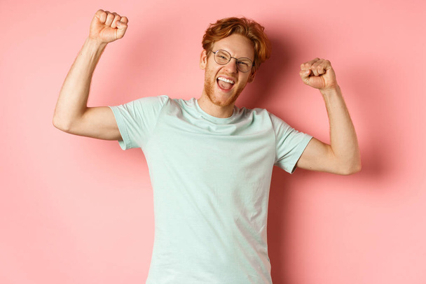 Fröhlicher junger Mann mit roten Haaren sieht glücklich aus, hebt die Hände in Pumps, feiert den Erfolg, fühlt sich wie ein Champion, gewinnt und steht vor rosa Hintergrund - Foto, Bild