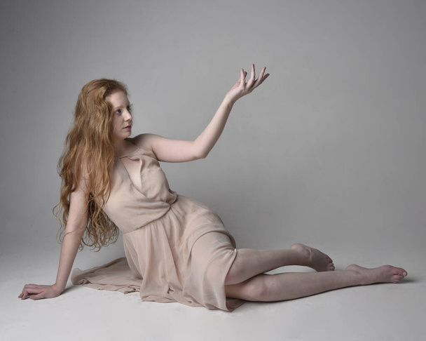 Full length portrait of pretty red haired woman dancer,  wearing skin toned flowing fairy dress. Sitting gestural poses  isolated on studio background. - Foto, afbeelding
