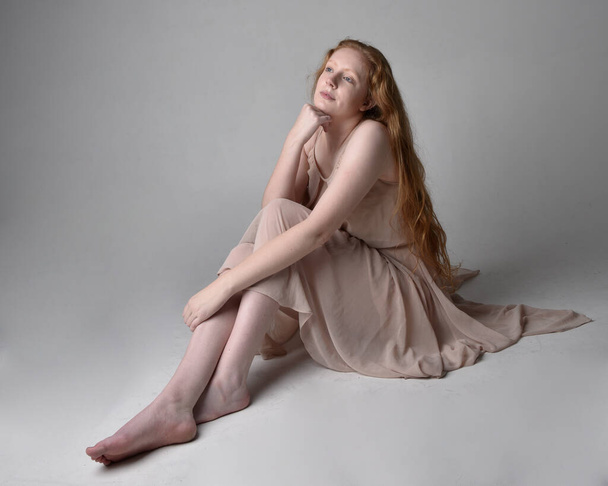 Full length portrait of pretty red haired woman dancer,  wearing skin toned flowing fairy dress. Sitting gestural poses  isolated on studio background. - Photo, Image
