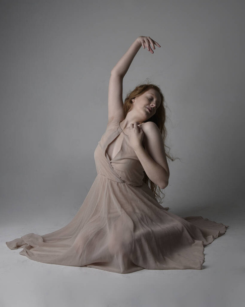 Full length portrait of pretty red haired woman dancer,  wearing skin toned flowing fairy dress. Sitting gestural poses  isolated on studio background. - Foto, afbeelding