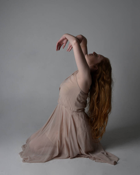 Полноразмерный портрет красивой рыжеволосой танцовщицы, одетой в тонированное сказочное платье. Сидя жесты позы изолированы на студийном фоне. - Фото, изображение