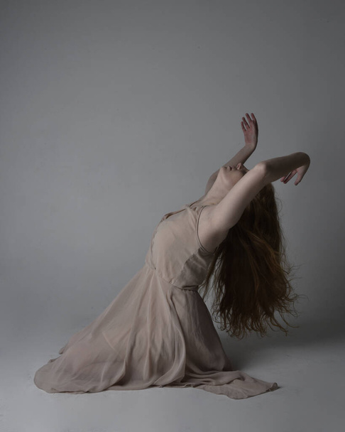 Full length portrait of pretty red haired woman dancer,  wearing skin toned flowing fairy dress. Sitting gestural poses  isolated on studio background. - Photo, Image