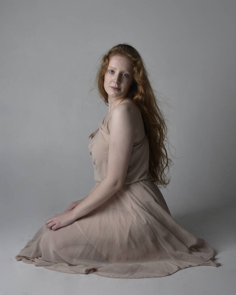 Full length portrait of pretty red haired woman dancer,  wearing skin toned flowing fairy dress. Sitting gestural poses  isolated on studio background. - Valokuva, kuva