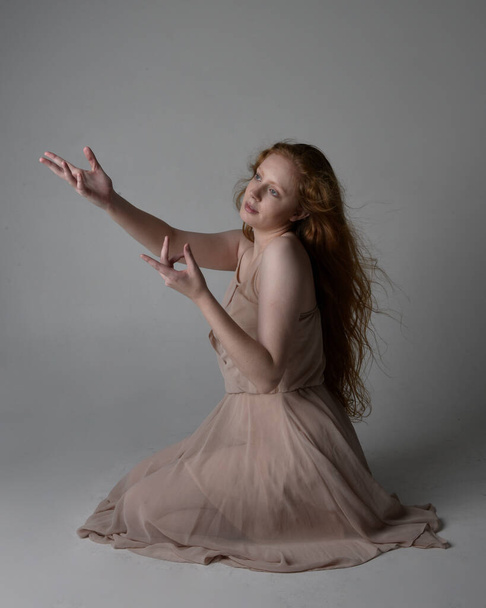 Full length portrait of pretty red haired woman dancer,  wearing skin toned flowing fairy dress. Sitting gestural poses  isolated on studio background. - Zdjęcie, obraz