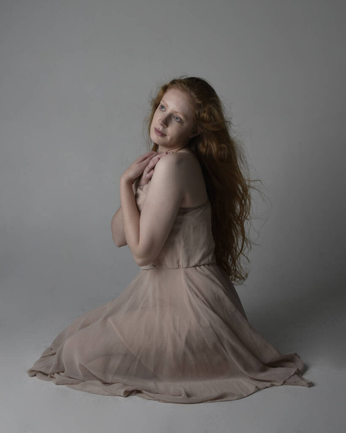 Portrait complet de jolie danseuse aux cheveux roux, vêtue d'une robe de fée fluide et tonique. poses gestuelles assis isolés sur fond de studio. - Photo, image