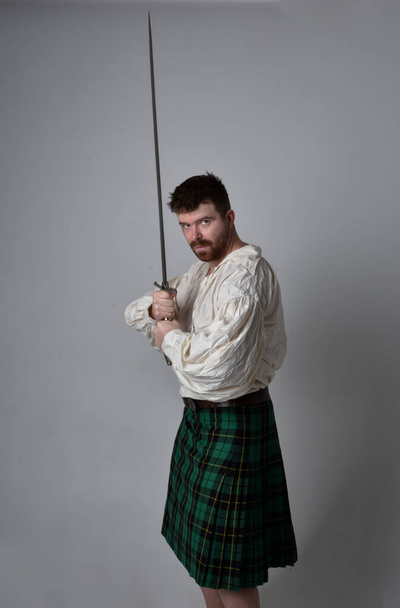 Zbliżenie portret przystojnego brunetka w szkockim kilcie i renesansowej białej bluzce pirata. Trzymając broń miecza, akcja poza izolacją na tle studia.    - Zdjęcie, obraz