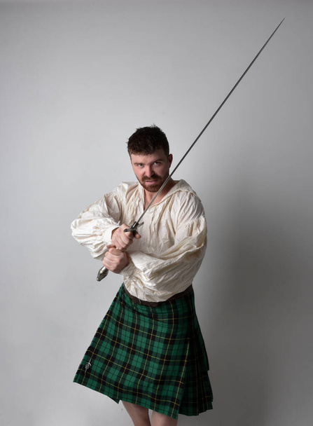 Close up portrait of handsome brunette man wearing Scottish kilt and renaissance white  pirate blouse shirt. Holding a sword weapon, action pose isolated against studio background.    - Foto, afbeelding