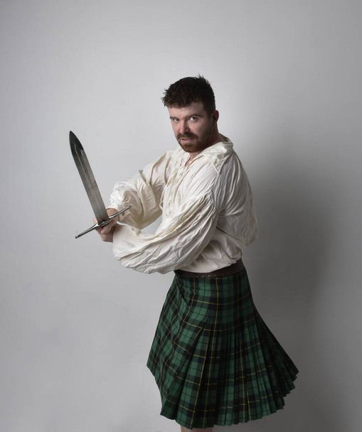 Κοντινό πορτραίτο ενός όμορφου μελαχρινού άντρα με σκωτσέζικη κιλτ και αναγεννησιακή άσπρη πειρατική μπλούζα. Κρατώντας ένα σπαθί όπλο, δράση θέτουν απομονωμένη σε στούντιο φόντο.    - Φωτογραφία, εικόνα
