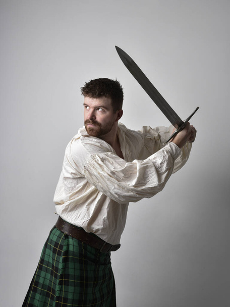 Close up portrait of handsome brunette man wearing Scottish kilt and renaissance white  pirate blouse shirt. Holding a sword weapon, action pose isolated against studio background.    - Photo, Image