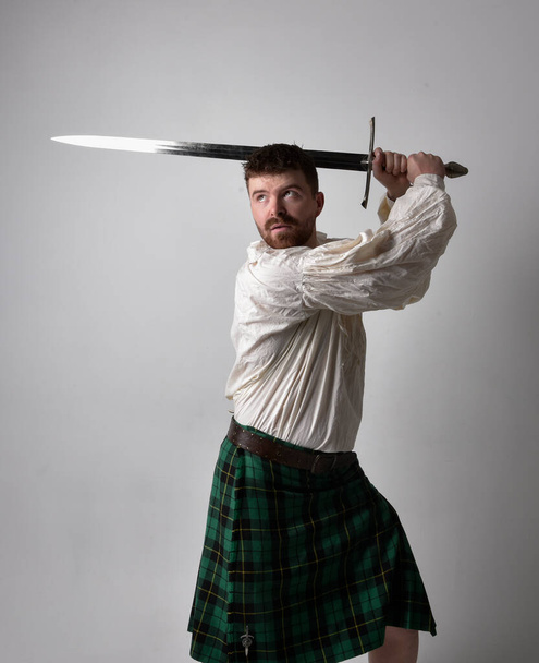 Close up portrait of handsome brunette man wearing Scottish kilt and renaissance white  pirate blouse shirt. Holding a sword weapon, action pose isolated against studio background.    - Foto, imagen