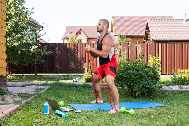 De sterke man gaat sporten in het park. Vrolijke sportieve man met zwart haar doet een squat met rubber en horloges in laptop in de tuin - Foto, afbeelding