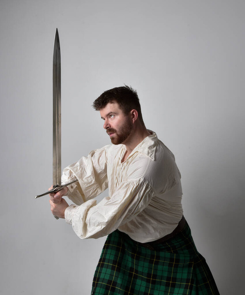 Close up portrait of handsome brunette man wearing Scottish kilt and renaissance white  pirate blouse shirt. Holding a sword weapon, action pose isolated against studio background.    - Фото, изображение