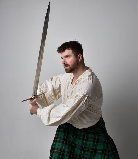 Zbliżenie portret przystojnego brunetka w szkockim kilcie i renesansowej białej bluzce pirata. Trzymając broń miecza, akcja poza izolacją na tle studia.    - Zdjęcie, obraz