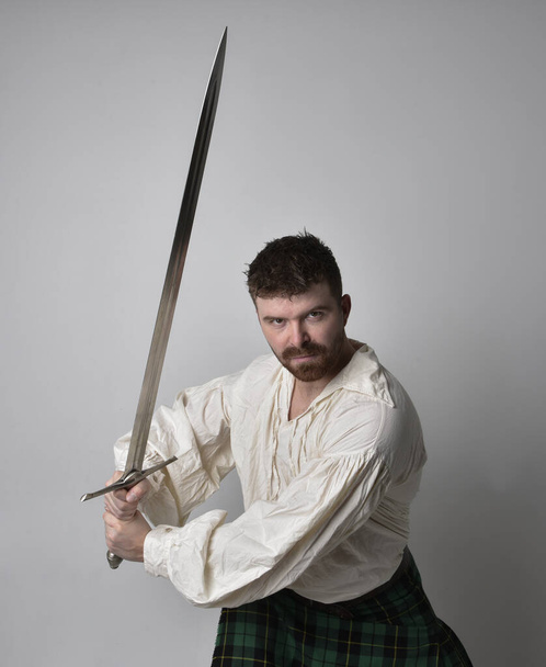 Gros plan portrait d'un bel homme brune portant un kilt écossais et un chemisier pirate blanc Renaissance. Tenant une épée, pose d'action isolée sur fond de studio.    - Photo, image