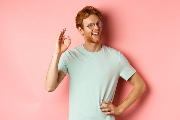 Cheerful guy with red hair and beard, wearing glasses, showing OK sign in approval and winking, smiling satisfied, standing over pink background - Photo, Image