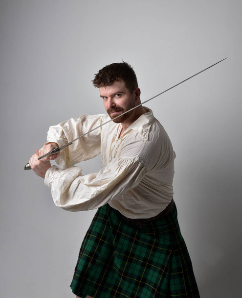 Close up portrait of handsome brunette man wearing Scottish kilt and renaissance white  pirate blouse shirt. Holding a sword weapon, action pose isolated against studio background.    - Photo, Image