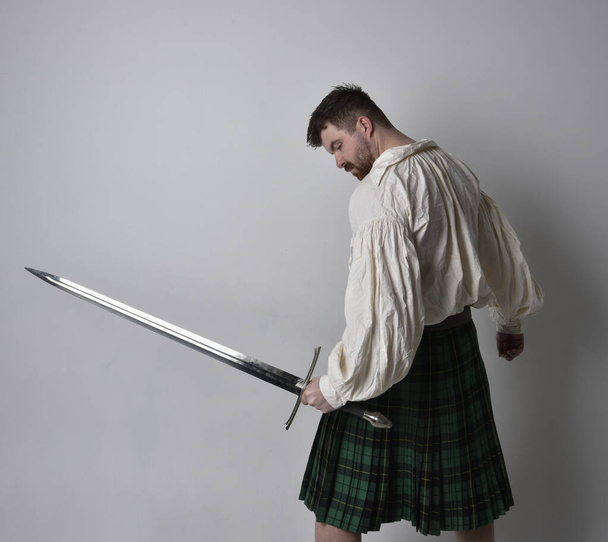 Close up portrait of handsome brunette man wearing Scottish kilt and renaissance white  pirate blouse shirt. Holding a sword weapon, action pose isolated against studio background.    - Φωτογραφία, εικόνα