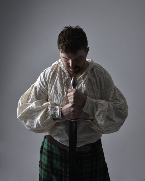 Close up retrato de homem morena bonito vestindo kilt escocês e renascimento blusa branca pirata. Segurando uma arma de espada, ação pose isolada contra o fundo do estúdio.    - Foto, Imagem