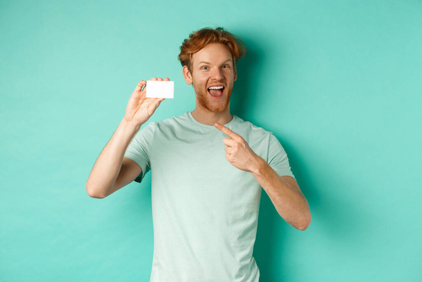 Σχέδιο αγορών. Όμορφος κοκκινομάλλης με t-shirt που δείχνει πλαστική πιστωτική κάρτα και χαμογελά, στέκεται πάνω από τυρκουάζ φόντο - Φωτογραφία, εικόνα