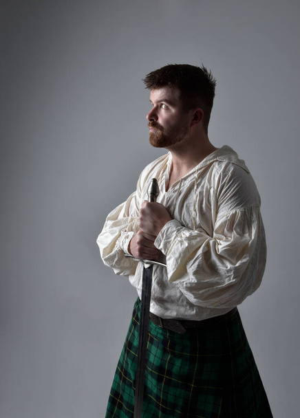 Κοντινό πορτραίτο ενός όμορφου μελαχρινού άντρα με σκωτσέζικη κιλτ και αναγεννησιακή άσπρη πειρατική μπλούζα. Κρατώντας ένα σπαθί όπλο, δράση θέτουν απομονωμένη σε στούντιο φόντο.    - Φωτογραφία, εικόνα