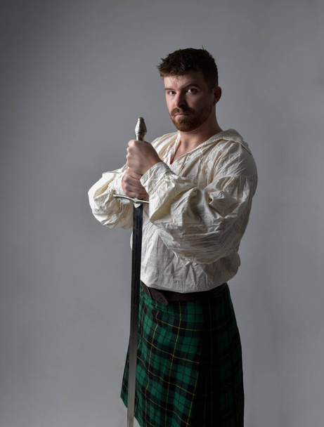 Close-up portret van knappe brunette man dragen Schotse kilt en renaissance witte piraat blouse shirt. Met een zwaardwapen, actie pose geïsoleerd tegen studio achtergrond.    - Foto, afbeelding