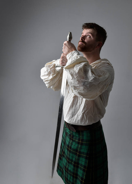Close-up portret van knappe brunette man dragen Schotse kilt en renaissance witte piraat blouse shirt. Met een zwaardwapen, actie pose geïsoleerd tegen studio achtergrond.    - Foto, afbeelding
