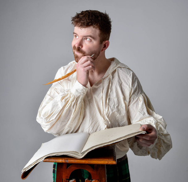Close up portrait of handsome brunette man wearing Scottish kilt and renaissance white  pirate blouse shirt. Holding a quill and writing in a book,  pose isolated against studio background.    - Foto, Imagen
