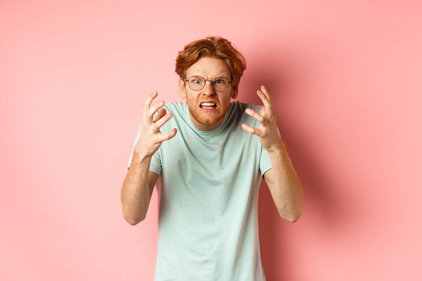Θυμωμένος κοκκινομάλλης με γυαλιά φωνάζοντας, συνοφρυωμένος και σφίγγοντας τα χέρια με απογοητευμένο και εξοργισμένο πρόσωπο, στέκεται πάνω από ροζ φόντο - Φωτογραφία, εικόνα