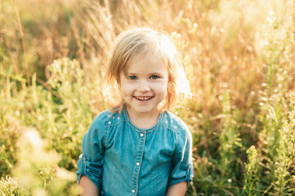 Close up portrait of happy cute little joyful blonde girl in denim dress in field of wildflowers at summer sunset. concept of joy of life, happiness. Sunny summer day at sunset. happy childhood - Photo, Image