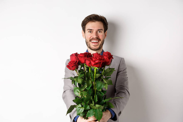 Красивый влюбленный мужчина желает счастливого Дня Святого Валентина, дарит букет цветов на романтическом свидании, улыбается в камеру, носит костюм на белом фоне - Фото, изображение