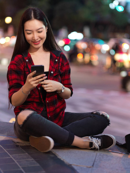 Porträt einer glücklichen Hipsterfrau, die auf einer Treppe sitzt und ihr Smartphone benutzt, mit Freunden chattet, Anwendungen herunterlädt, soziale Medien aktualisiert, verschwommene Nachtlichter im Hintergrund - Foto, Bild